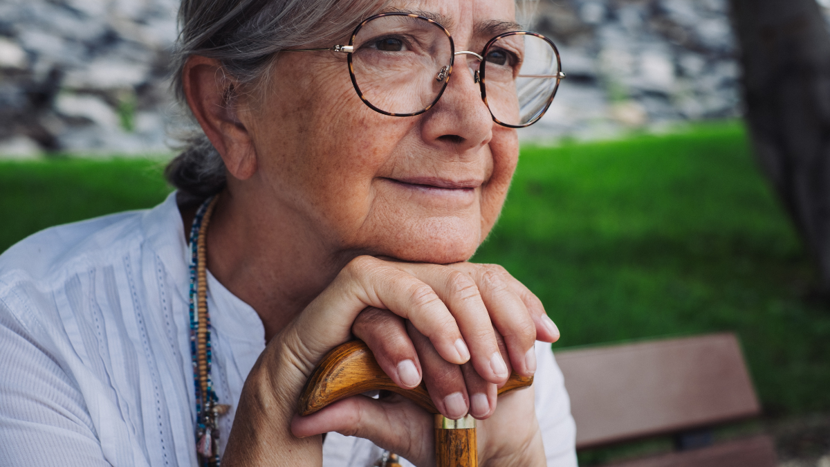 Portrait of a thoughtful elderly woman of 70 years old sitting on a bench in the park leaning on a stick due to walking disability, the elderly lady looks away.
