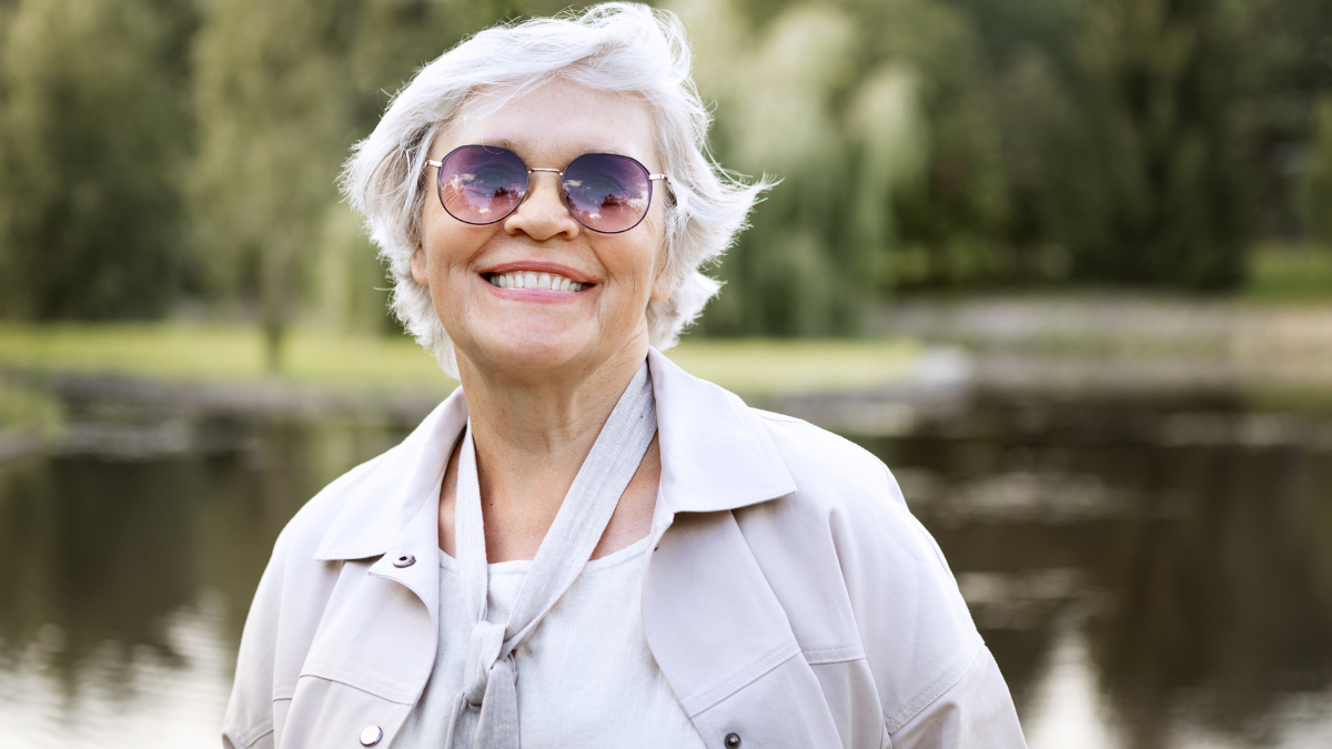 Happy senior woman with white hair smiling outdoors, wearing sunglasses and jacket, enjoying active retirement lifestyle, positive aging, freedom, joy, wellness and happiness in nature park background.