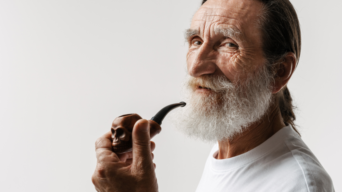 European senior man looking at camera while posing with smoking pipe isolated over white wall.