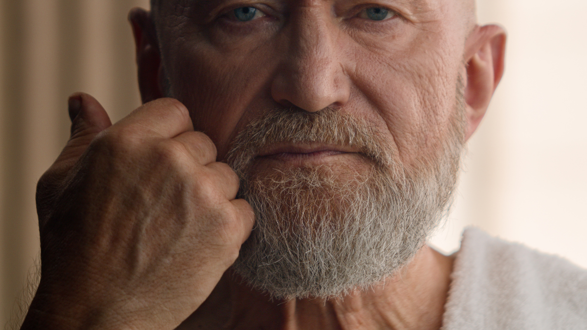 Caucasian old retired bearded man elderly male 60s person bearded senior grandfather touch beard guy mature touching face gray hair skin care portrait close up with bath towel on shoulder in bathroom.