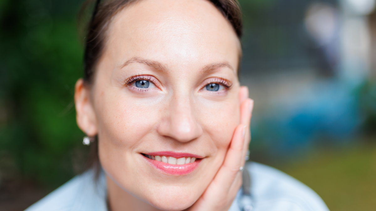 Close-up detail portrait view of young adult beautiful woman holding head chin with hand relaxed smiling sitting outdoors on day light. Female person close natural calm emotions outside.
