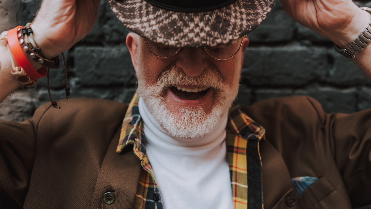Close up portrait of happy pensioner smiling and taking off his hat near black brick wall.