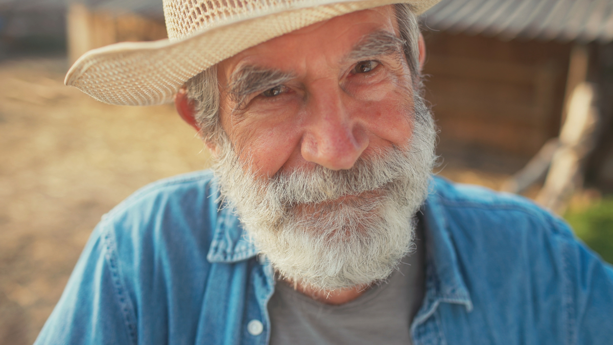 Close portrait view of older Caucasian male with grey beard and moustache. Man looking directly at camera while smiling. Farmer proud of his farm. Wearing yellow straw had and blue shirt.
