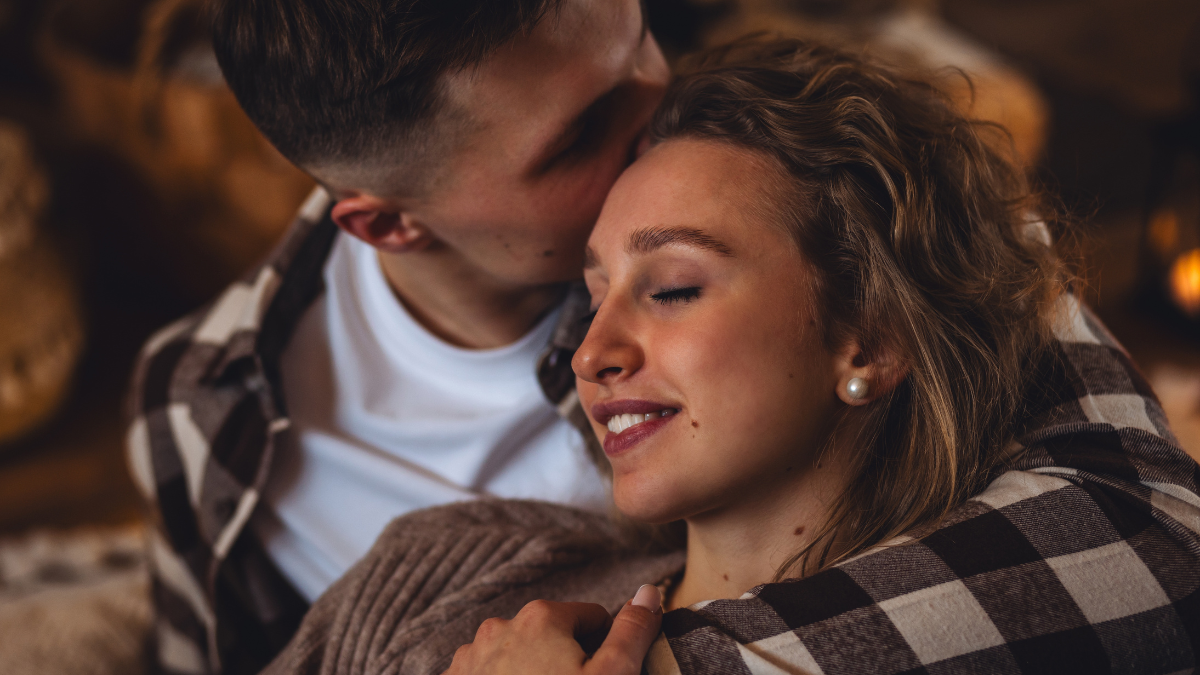 Beautiful young loving couple relaxing near fireplace in bed at home. Happy spouses enjoying lazy romantic winter cozy morning in bedroom. Candles, garlands, countryside interior. Romantic date