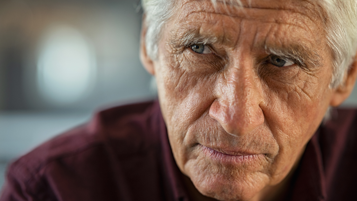 Thoughtful senior man sitting on couch looking away with copy space. Depressed sad man thinking at home. Elderly pensive senior suffering from alzheimer disease.