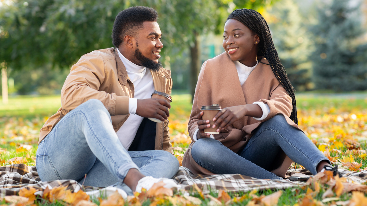 Outdoor date ideas concept. Smiling african american couple drinking takeout coffee while sitting on picnic in autumn park.