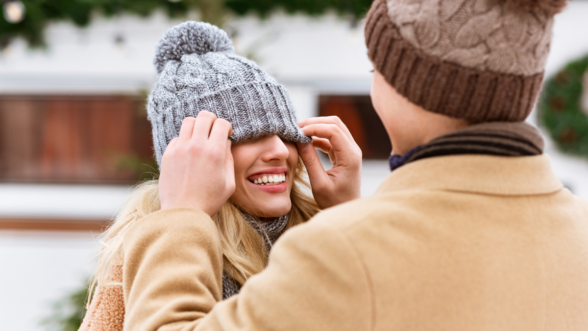 Romantic couple having fun outdoors in winter day, loving man playing with girlfriend's hat, covering her eyes and having Low-Cost Winter Date Ideas.