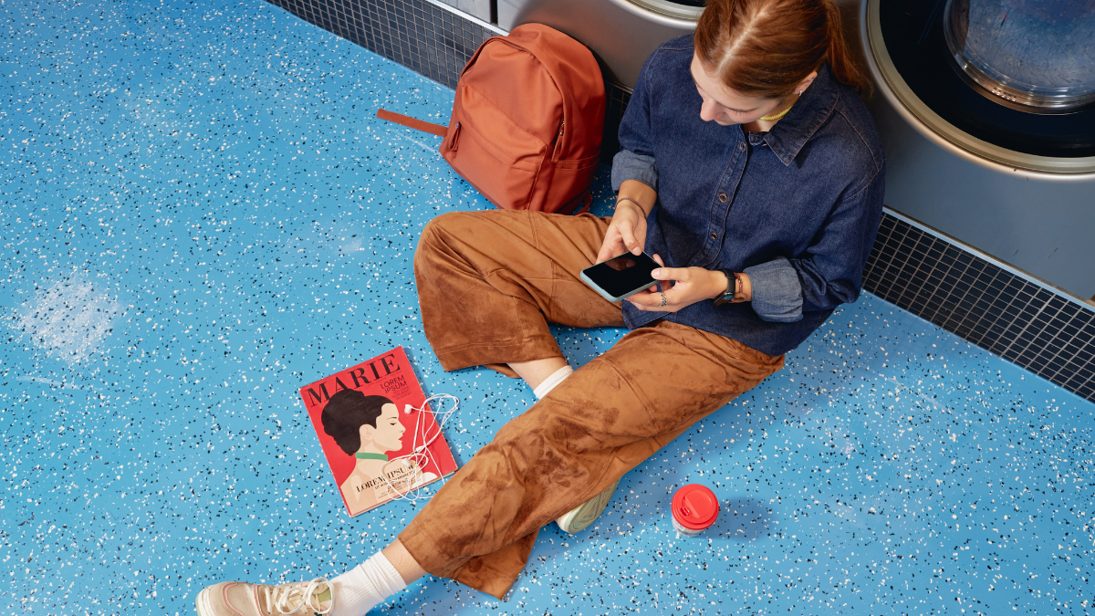 Top view of young woman text messaging on phone sitting on floor while passing time in self service laundry waiting for clean clothes, concept frugal living rules for everyday life.