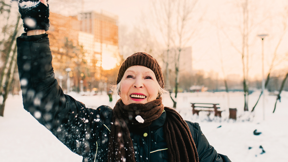senior woman in hat and sporty jacket snowballing in snow winter park. Winter, age, sport, activity, season concept.