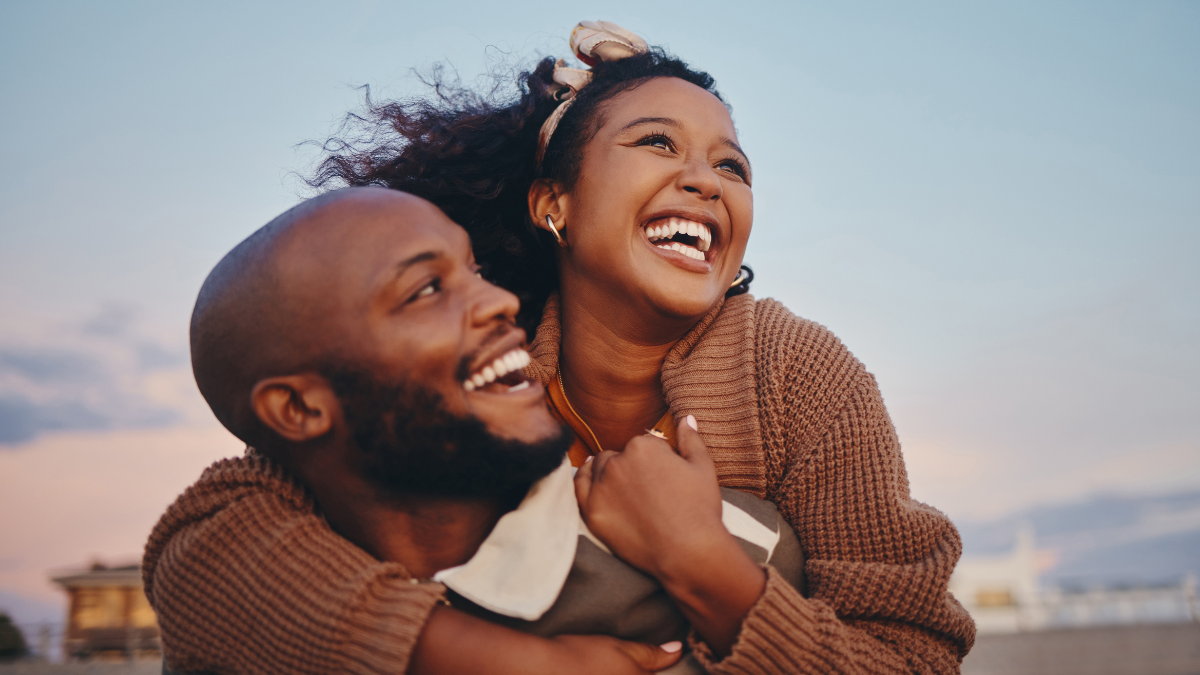 Happy black couple, love and hug laughing in joyful happiness for bonding time together in the outdoors. African American man and woman enjoying playful fun piggyback with smile for holiday in nature.