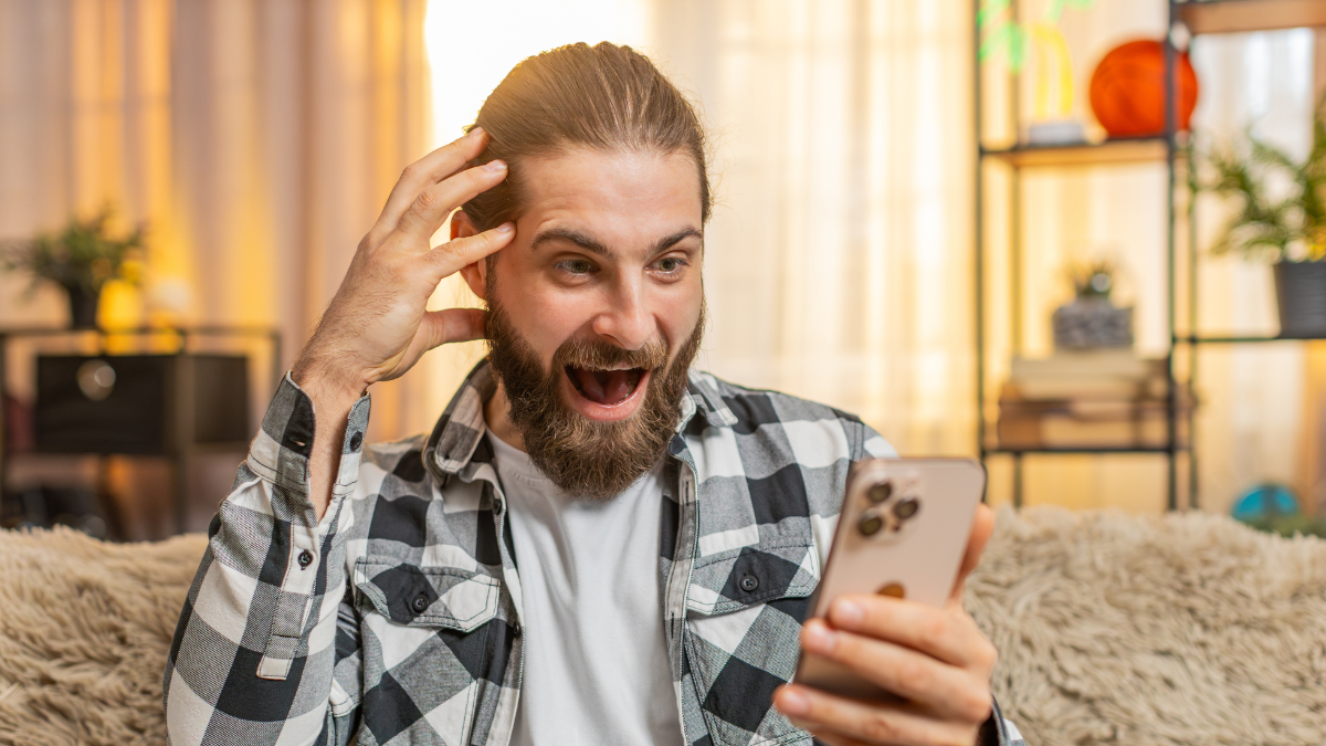 Young man sits at home rapidly inputting text on phone, seeking perfect discount in online store market place. Guy sitting on sofa exclaiming wow expression while discovering a highly beneficial offer how to save money fast.