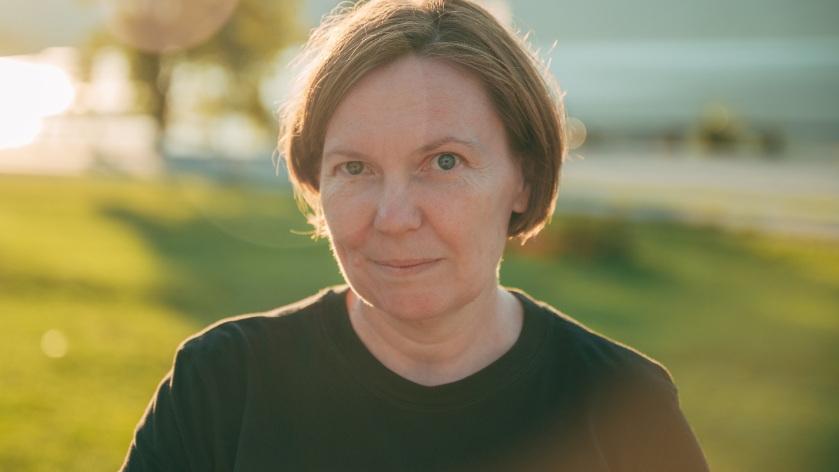Outdoor portrait of mature brunette woman in park at golden sunset, looking at camera with intriguing smile, soft lens flare creating elegant and timeless mood. Selective focus.
