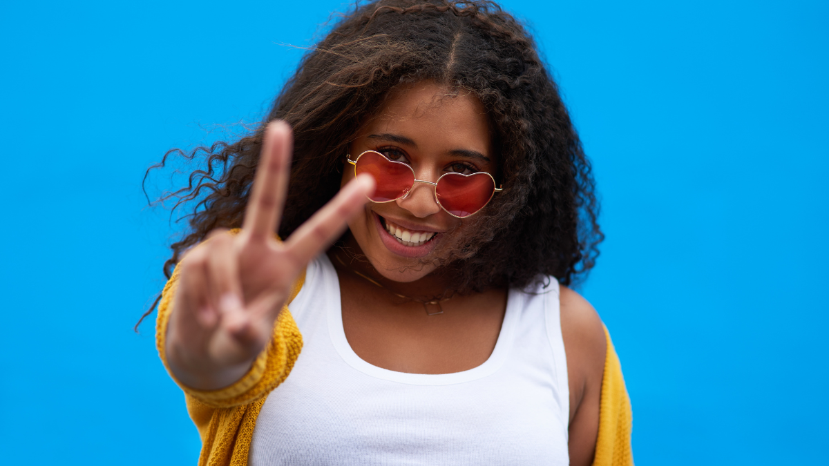 Portrait, woman and happy with peace sign in studio for gen z fashion, heart glasses and aesthetic style. Excited, girl and cool v icon with trendy eyewear, creative and opinion on blue background.