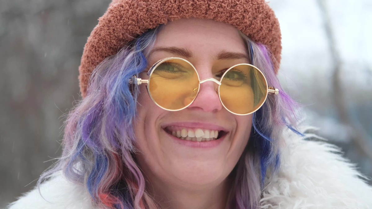 Portrait of a woman in sunglasses and a white fur coat in winter. Woman with colored curly hair posing outdoors during snowfall.