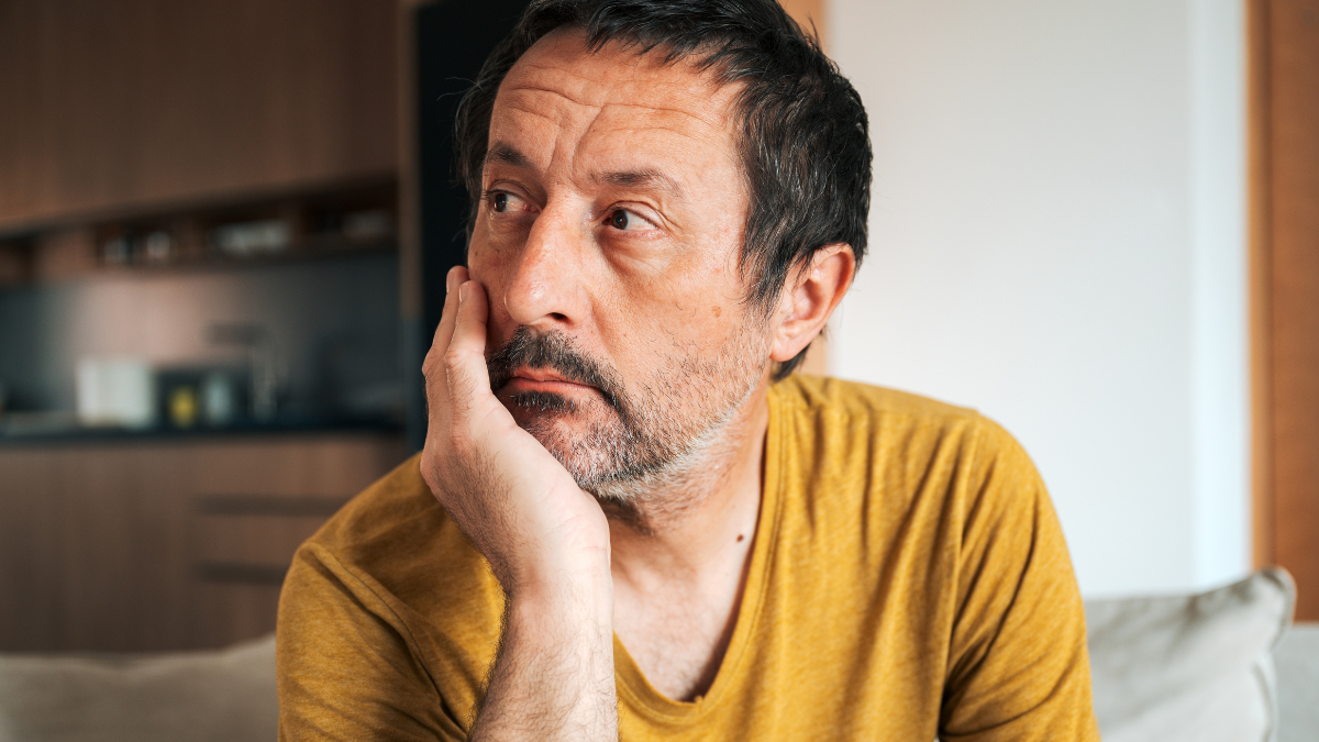 Bored middle-aged man sits on a sofa at home, resting his face on his hand and staring into the distance. Selective focus.
