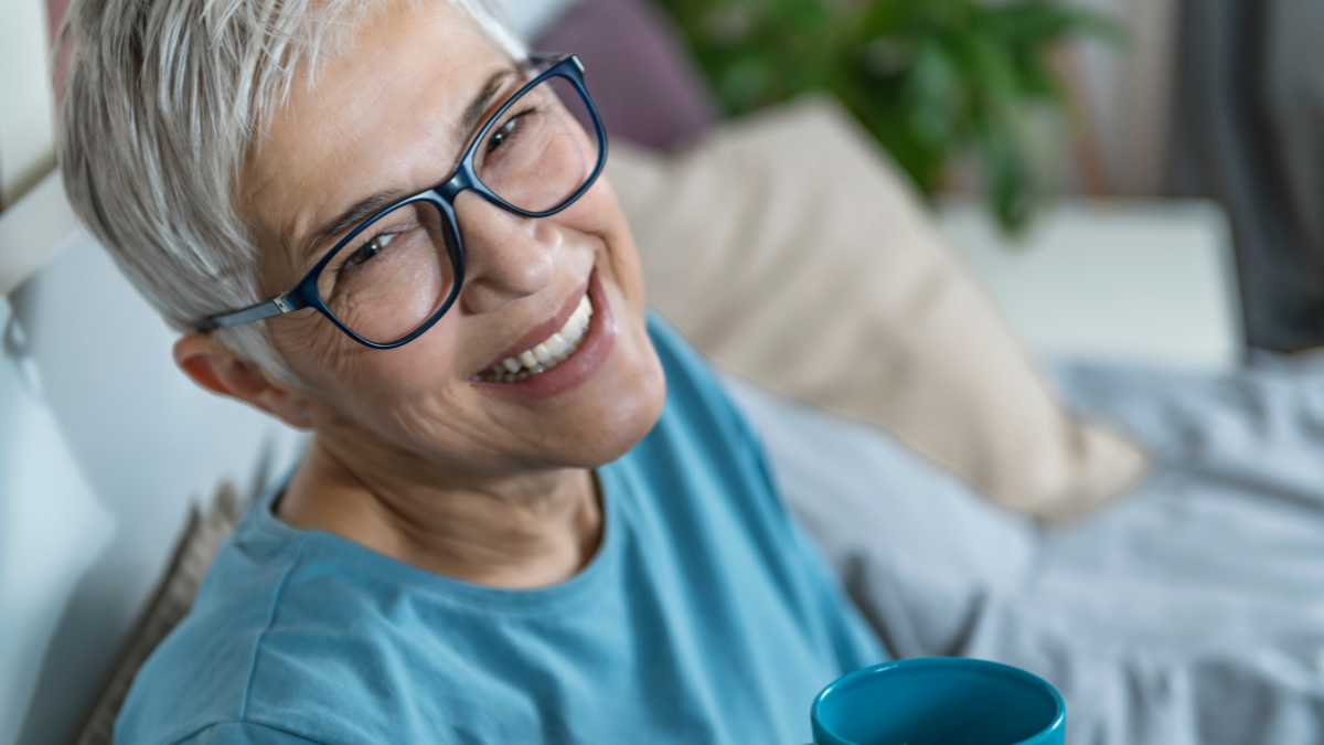 Smiling mature woman lying in bed, drinking coffee and talking over the phone before sleep New Frugal Habits Boomers Are Adopting to Survive Retirement.