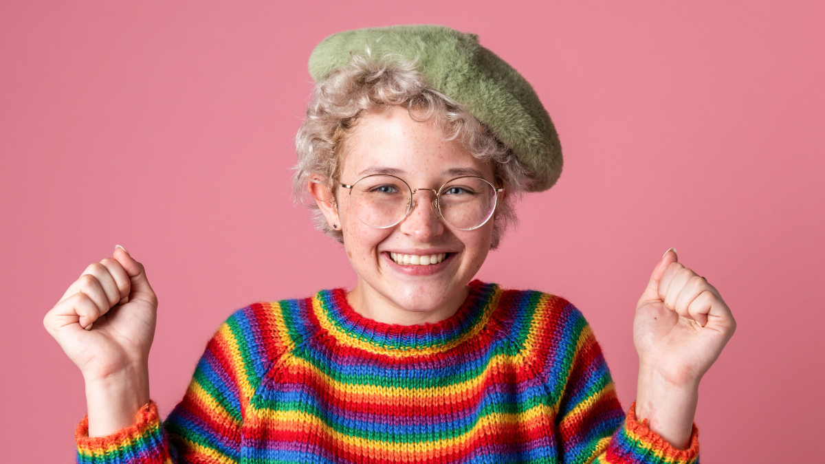 A person with curly hair, wearing glasses and a green beret, smiles joyfully. They are dressed in a colorful rainbow sweater against a pink background free things to do.