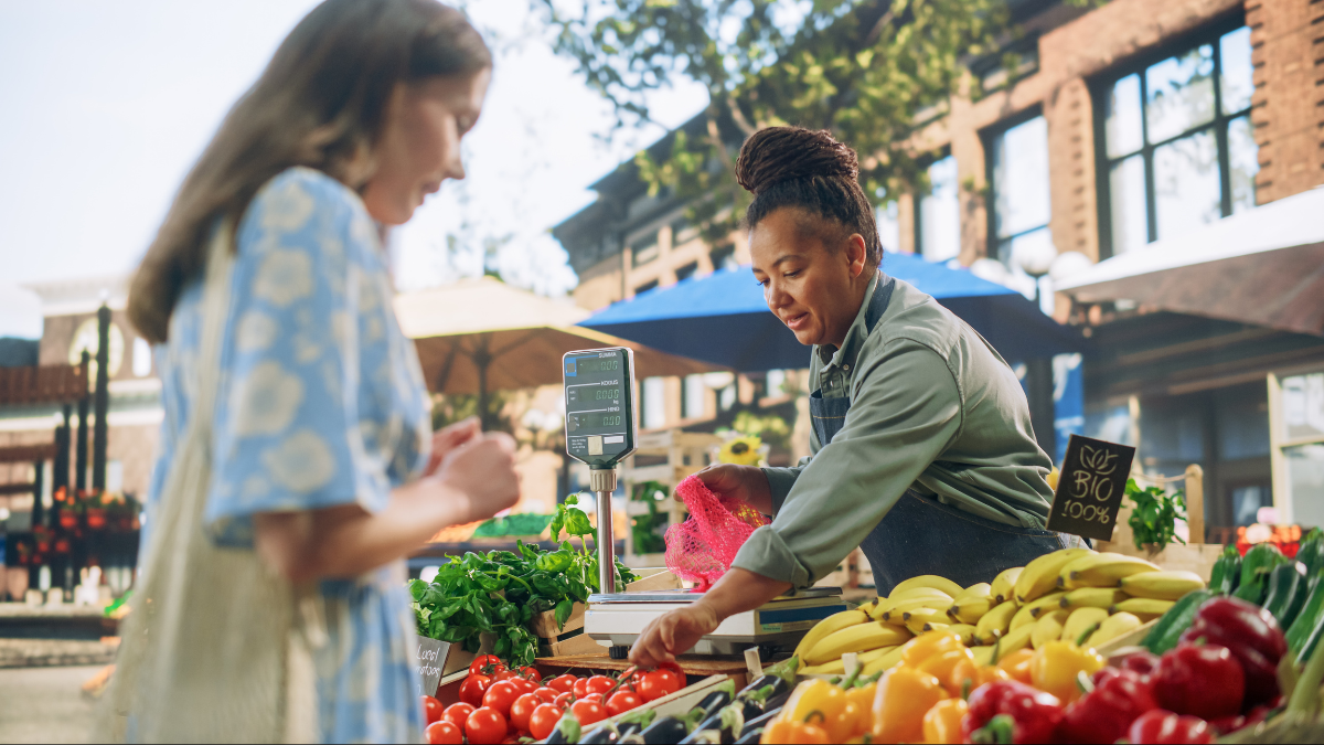Middle Aged Black Female Running a Small Business on a Farmers Market, Selling Organic Fruits and Vegetables. Young Female Shopping For Food, Choosing a Fresh Courgette or Zucchini lower your monthly expenses.
