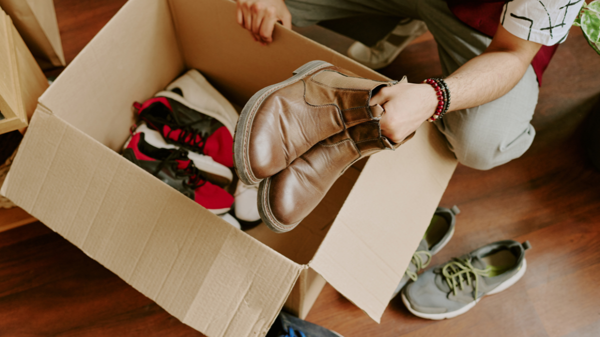 Person sorting various pairs of shoes in cardboard box on wooden floor. Different styles of shoes present, with some already inside box and others around it on floor.
