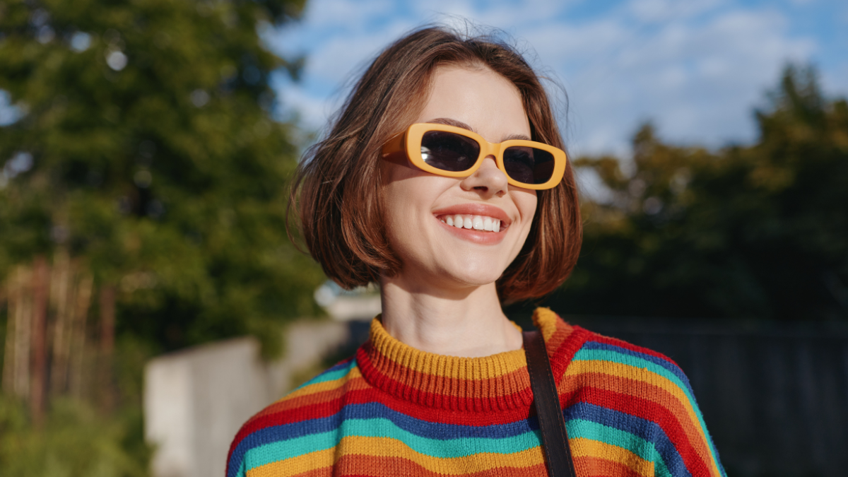 Woman in her late 20s with short hair smiling while walking outdoors in sunny park, wearing rainbow striped sweater and yellow sunglasses, casual fashion lifestyle portrait.