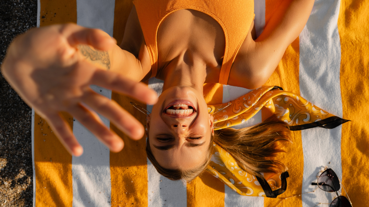 young attractive smiling happy blond woman lying on striped blanket in sun in yellow swimsuit tanning on summer vacation, season fashion trend accessories, top view from above, teeth smile, beach sea. Fun Things To Do This Weekend.