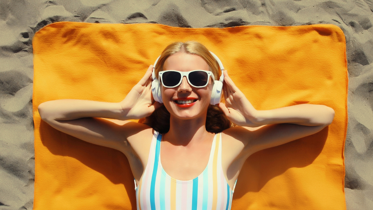 Summer vacation, tourism, happy cheerful young woman listening to music lying on sand on the beach, relaxed tourist girl in headphones on towel in swimsuit, glasses, sunny day, top view. save money this summer.